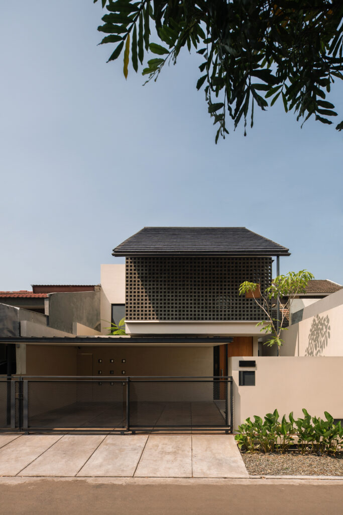 Front view of A&T Residence, a two-story modern tropical house with a clean beige façade, gable roof, and prominent upper-level square concrete vent block screen, constructed by NDA in collaboration with architect Dhaniesal.